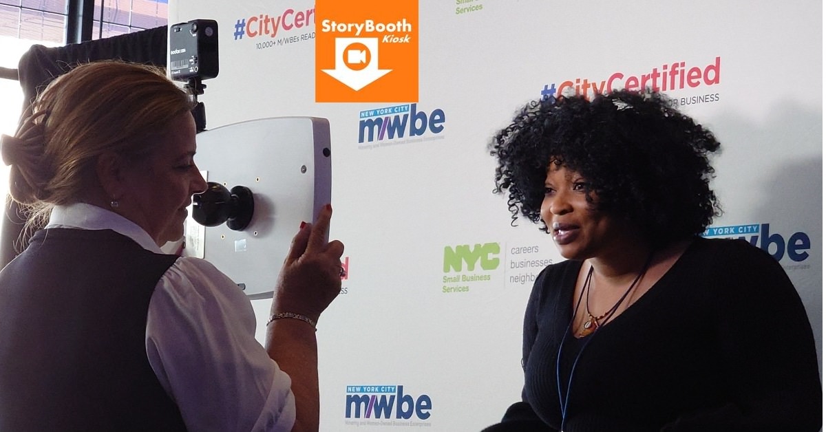 A StoryBooth Kiosk host holds up a roaming StoryBooth Kiosk Pro for a female to record her video in front of a backdrop featuring logos of NYC Small Business Services